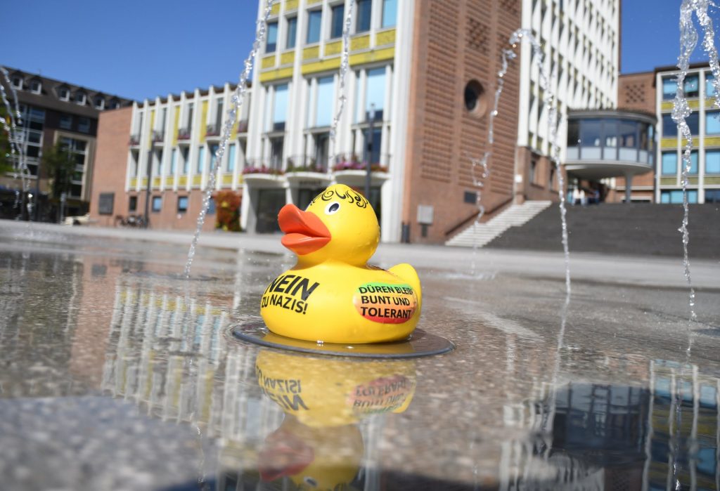 Die bemalte Ente steht auf den Wasserspielen Kaiserplatz, im Hintergrund das Rathaus.