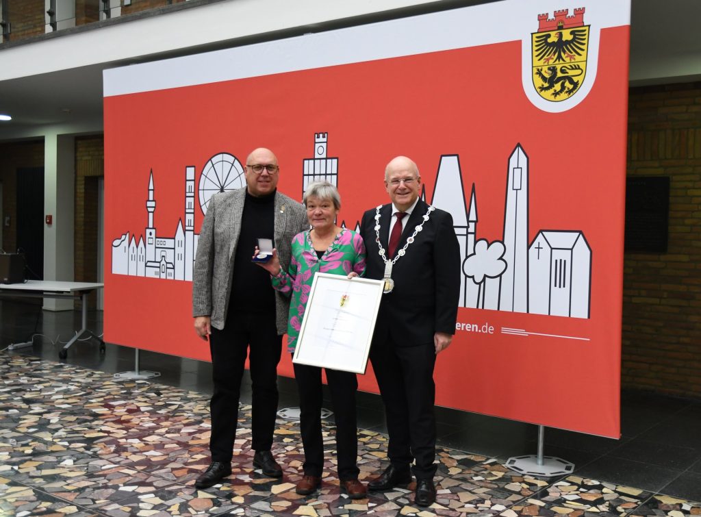 Das Foto zeigt Dirk Nieveler, Verena Schloemer mit Medaille und Urkund sowie Bürgermeister Frank Peter Ullrich vor der Skyline von Düren.