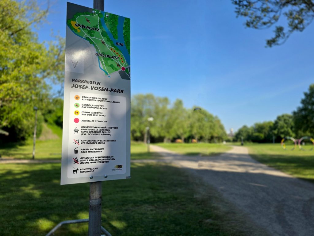Schild mit den Parkregeln des Josef-Vosen-Parks und dem Park im Hintergrund