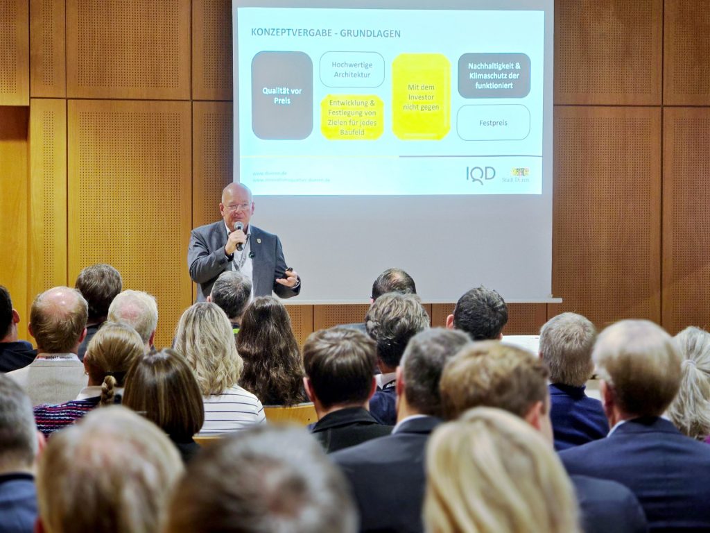 Mann steht vor Publikum und spricht in ein Mikrofon