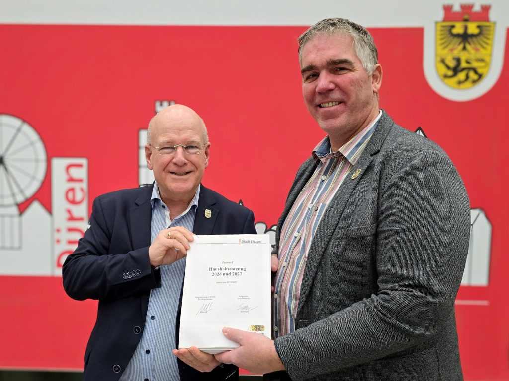 Zwei Männer halten einen großen Stapel gebundener Blätter in die Kamera