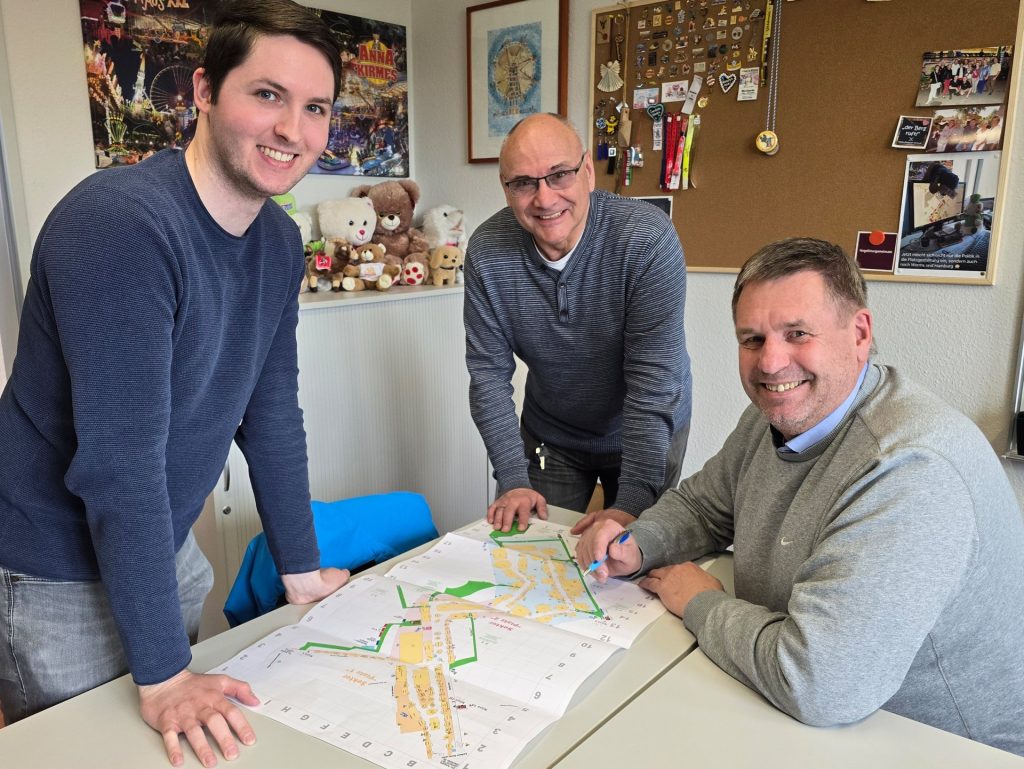 drei Männer stehen um eine Tisch herum, auf dem Tisch liegt ein Plan