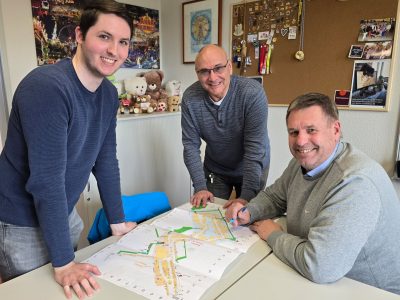 drei Männer stehen um eine Tisch herum, auf dem Tisch liegt ein Plan
