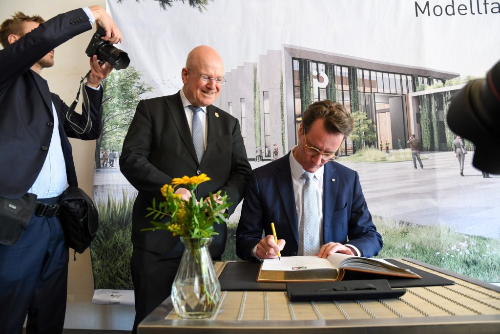 Im Beisein von Bürgermeister Frank Peter Ullrich trägt sich Ministerpräsident Hendrik Wüst (sitzend) in das Goldene Buch der Stadt Düren ein. Im Hintergruund ist eine Leinwand, auf der das Modell des IQD zu sehen ist.