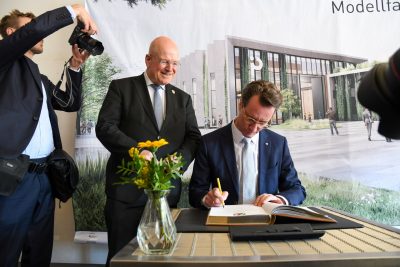 Im Beisein von Bürgermeister Frank Peter Ullrich trägt sich Ministerpräsident Hendrik Wüst (sitzend) in das Goldene Buch der Stadt Düren ein. Im Hintergruund ist eine Leinwand, auf der das Modell des IQD zu sehen ist.