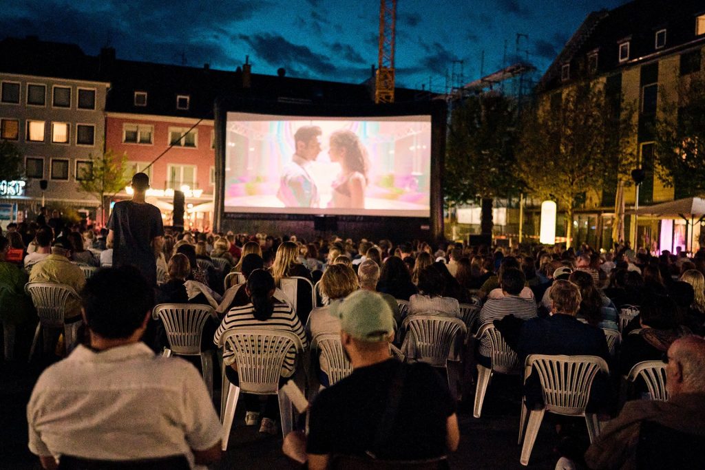Das Foto zeigt die große Open-Air-Kinoleinwand auf dem Marktplatz. Davor sitzen Leute.